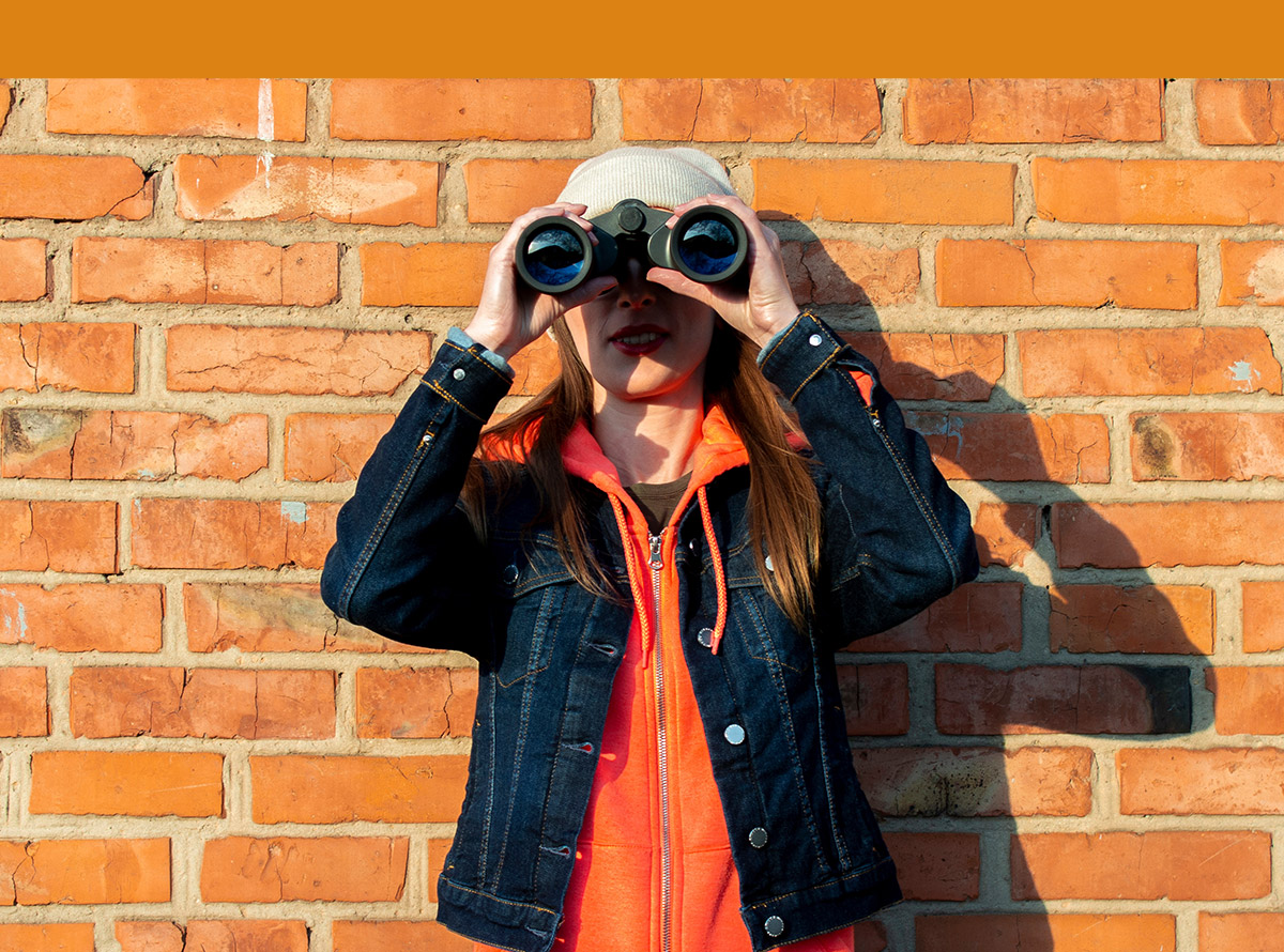 Young woman looking through binoculars on the background of a brick wall, wearing a gray cap and a denim jacket